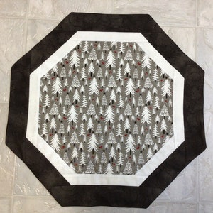 May include: Octagonal table runner with a white and brown geometric design. The center features a repeating pattern of white stylized trees on a gray background with red berries.
