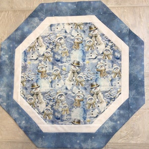 May include: An octagonal quilted table topper featuring a winter scene with snowmen. The central design shows snowmen wearing hats and scarves against a snowy backdrop. The topper has a white border and a blue snowflake patterned edge.