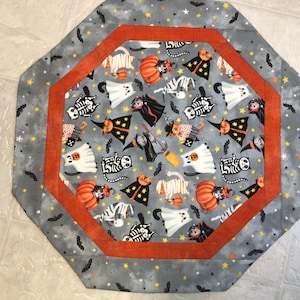 May include: Octagonal Halloween-themed table runner. The gray fabric features a pattern of ghosts, cats, skeletons, and pumpkins. An orange border frames the central design, with a gray border featuring bats and stars.