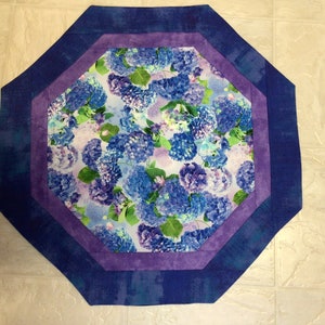 May include: Octagonal table runner with a floral pattern featuring blue and purple hydrangeas. The runner has a multi-layered border in shades of blue and purple.