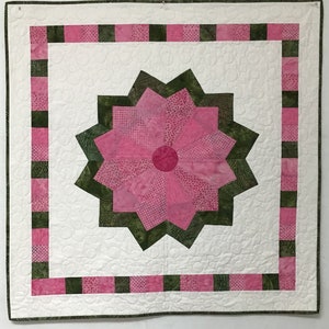 May include: A white quilt with a pink and green geometric design. The center of the quilt features a large pink flower with a green border. The quilt is edged with a pink and green border.