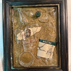 May include: A framed mixed-media artwork featuring fragments of a Buffalo Trace bourbon bottle, label, and cap. The artwork has a dark wooden frame and a textured, golden-brown background. The bottle fragments are clear, and the label is tan with black text.