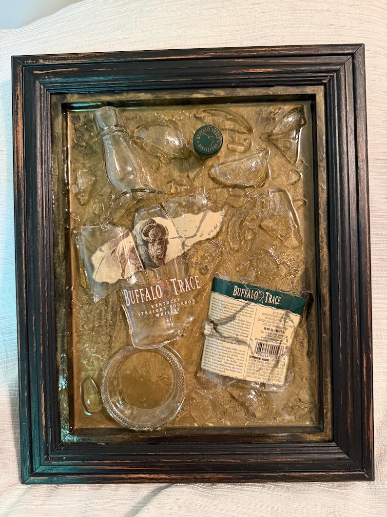 May include: A framed mixed-media artwork featuring fragments of a Buffalo Trace bourbon bottle, label, and cap. The artwork has a dark wooden frame and a textured, golden-brown background. The bottle fragments are clear, and the label is tan with black text.