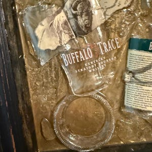 May include: Artwork featuring fragments of a Buffalo Trace Kentucky Straight Bourbon Whiskey bottle. The image includes the bottle's label with a bison illustration and text, along with broken glass pieces, all set within a dark wooden frame.