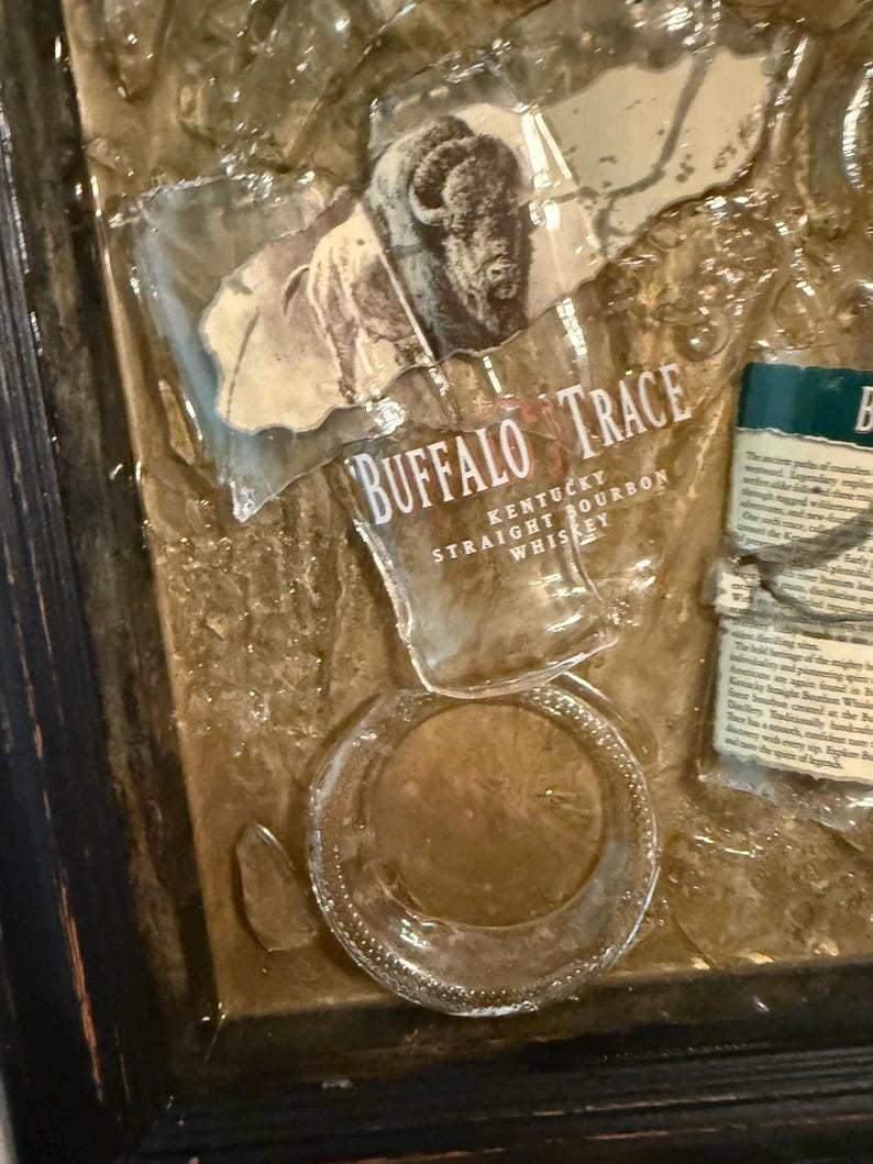 May include: Artwork featuring fragments of a Buffalo Trace Kentucky Straight Bourbon Whiskey bottle. The image includes the bottle's label with a bison illustration and text, along with broken glass pieces, all set within a dark wooden frame.