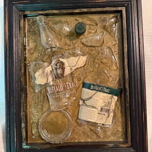 May include: A framed mixed-media artwork featuring fragments of a Buffalo Trace whiskey bottle, label, and cap. The artwork has a dark brown frame and a textured, golden-brown background. The label reads "Buffalo Trace Kentucky Straight Bourbon Whiskey."