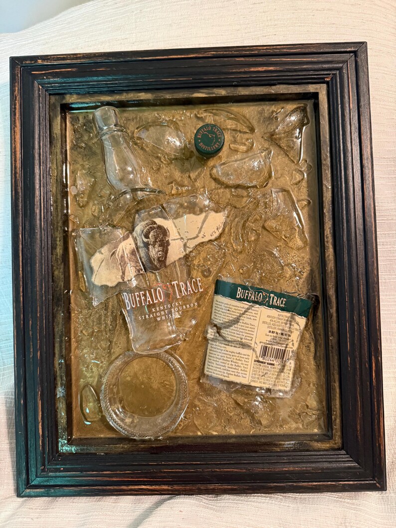 May include: A framed mixed-media artwork featuring fragments of a Buffalo Trace whiskey bottle, label, and cap. The artwork has a dark brown frame and a textured, golden-brown background. The label reads "Buffalo Trace Kentucky Straight Bourbon Whiskey."