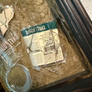 May include: Artwork featuring fragments of a Buffalo Trace bourbon bottle label and glass, set against a textured, brown background. The label, with green and white text, is partially torn. The frame is dark wood.