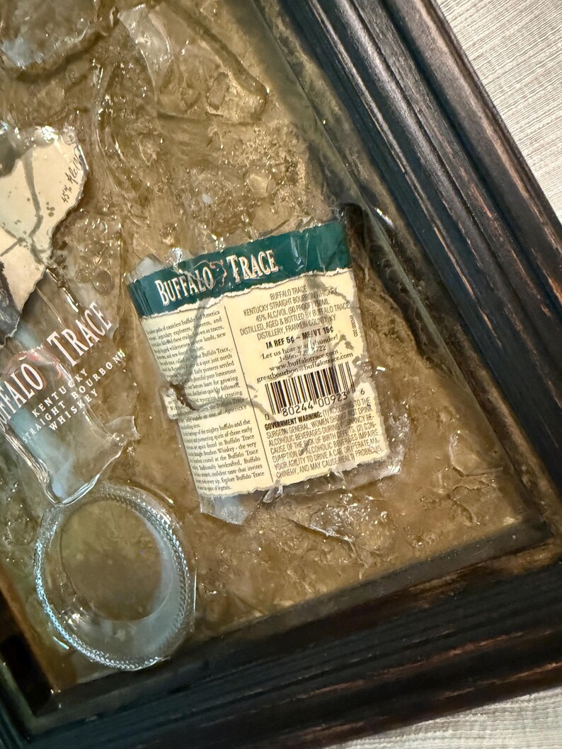 May include: Artwork featuring fragments of a Buffalo Trace bourbon bottle label and glass, set against a textured, brown background. The label, with green and white text, is partially torn. The frame is dark wood.