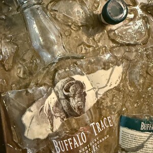 May include: A clear glass bottle and a green-topped cap are set against a textured, golden-brown background. The label for "Buffalo Trace Kentucky Straight Bourbon Whiskey" is visible, featuring a bison illustration.