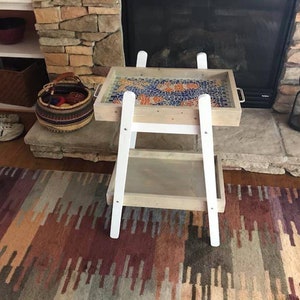 May include: White serving cart with two shelves. The top shelf has a wooden tray with a colorful mosaic tile design. The cart is in front of a stone fireplace.