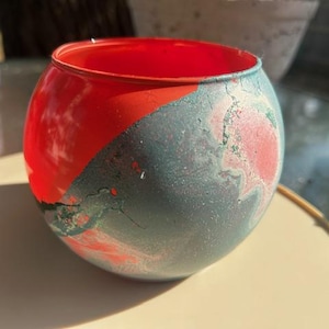 May include: A round, hand-painted glass bowl with a vibrant red interior and a marbled exterior in shades of teal, gray, and red. The bowl is sitting on a light-colored surface, showcasing its unique color palette and design.