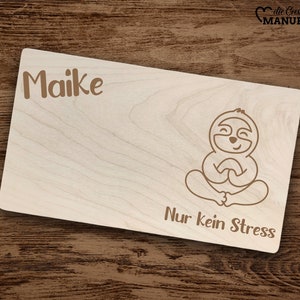 Può includere: Un cartello di legno con il nome "Maike" e un cartone animato di un bradipo seduto in una posa yoga. Il cartello recita "Nur kein Stress", che si traduce in "Solo niente stress" in italiano.