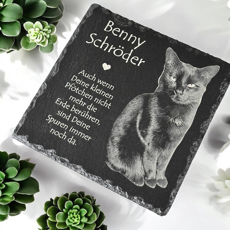 Puede incluir: Placa conmemorativa de pizarra negra con una imagen grabada de un gato. La placa incluye el nombre "Benny Schr&ouml;der" y un texto en alem&aacute;n. El gato est&aacute; representado en posici&oacute;n sentada.