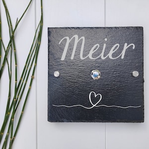 Könnte beinhalten: Schwarzes Schiefer-Schild mit dem Namen "Meier" in weißer, kursiv geschriebener Schrift. Das Schild hat ein kleines Herz und eine Wellenlinie unter dem Namen. Es befinden sich zwei silberne Türklingeln auf dem Schild.