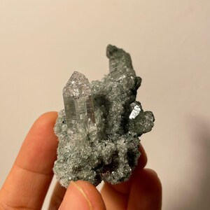 Może przedstawiać: Gromada przezroczystych kryształ&oacute;w kwarcu z zieloną, mchową macierzą. Kryształy są spiczaste i mają szklisty wygląd.
