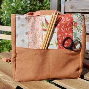 Könnte beinhalten: Eine pfirsichfarbene Tragetasche mit Patchwork-Design. Die Tasche hat eine Vordertasche und ist mit Stricknadeln aus Holz und einer schwarzen Schere gefüllt. Der Patchwork-Stoff weist Blumen- und geometrische Muster in Pfirsich-, Weiß- und Grüntönen auf.