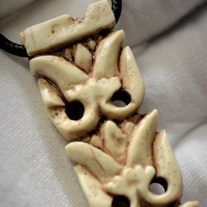 May include: A cream-colored pendant necklace with a detailed carved design of three stylized flowers. The pendant is attached to a black cord.