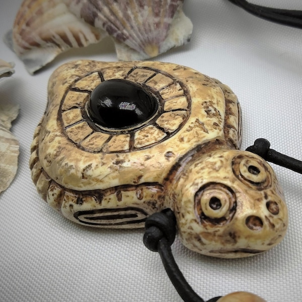 May include: A beige ceramic turtle pendant with a black circular inlay, hanging from a black leather cord with wooden beads. 