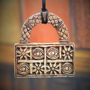 Puede incluir: Un colgante blanco con un diseño marrón que presenta ojos y flores. El colgante está unido a un cordón negro.