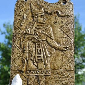 Può includere: Una placca in bronzo che raffigura una figura con un cappello alto e abiti elaborati. La figura si trova su una superficie con motivi, simboli e disegni. La placca ha una superficie testurizzata e una forma leggermente curva.