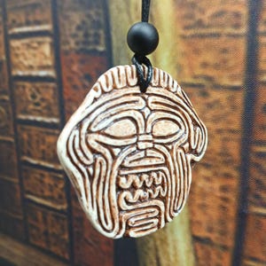 May include: A pendant necklace with a carved tribal mask design in off-white and brown. The pendant hangs from a black cord with a black bead. The mask features detailed lines and a stylized face.