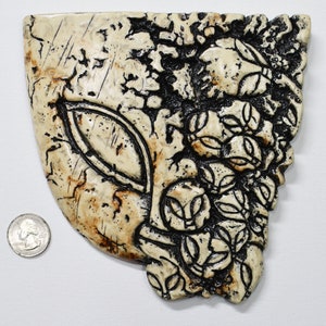 Puede incluir: Una escultura de pared de cerámica con forma de rostro con un acabado agrietado y desgastado. El rostro está pintado en tonos beige y marrón con acentos negros. La escultura mide aproximadamente 15 cm de alto y 13 cm de ancho.