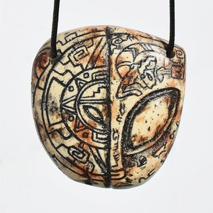 Può includere: Una collana con un ciondolo in ceramica e un cordoncino nero. Il ciondolo è color crema con segni marroni e neri. Il ciondolo ha un design geometrico su un lato e un viso stilizzato sull'altro.