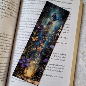 K&ouml;nnte beinhalten: Dekorativer Lesezeichen mit einer fantastischen Illustration eines Glases voller Blumen und Schmetterlinge. Das Kunstwerk hat eine dunkle, vertr&auml;umte &Auml;sthetik mit Farbt&ouml;nen von Lila, Blau und Gold. Das Lesezeichen befindet sich in einem offenen Buch.