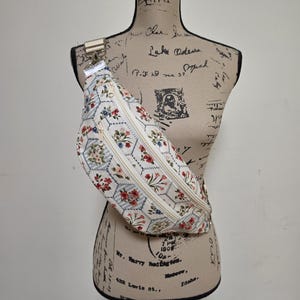 May include: A fabric fanny pack featuring a cream-colored base with a repeating pattern of blue hexagonal outlines, small red and blue floral clusters, and thin blue stripes. The bag includes two front zippered compartments with cream-colored zippers.
