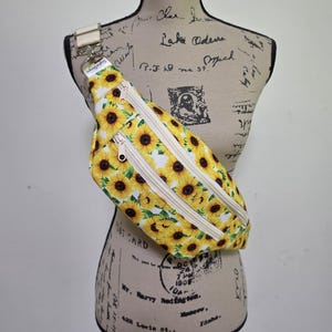 May include: A crossbody fanny pack featuring a vibrant sunflower pattern on a white background. The bag includes two front zippered compartments with light-colored zippers and an adjustable strap attached to a metal ring with a small white label.