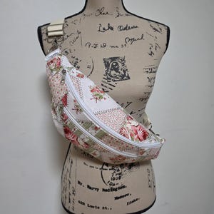 May include: A crossbody fanny pack featuring a patchwork design with floral patterns in shades of pink, green, and cream. The bag includes two white zippered compartments, a white adjustable strap with silver hardware, and a small white brand label.