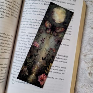 K&ouml;nnte beinhalten: Rechteckiger Lesezeichen mit einer fantastischen Illustration einer Motte, Blumen und einem Vollmond. Das Kunstwerk ist vor einem dunklen, vertr&auml;umten Hintergrund mit einem Hauch von Glanz platziert. Das Lesezeichen liegt auf einem offenen Buch.