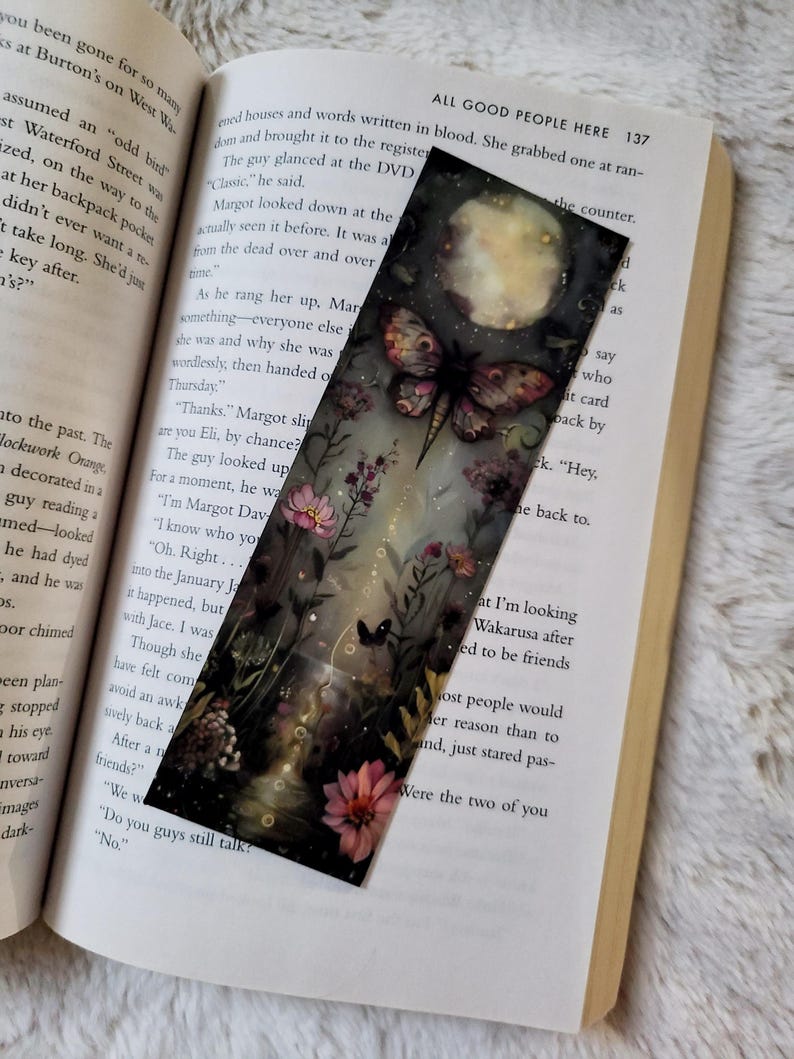 K&ouml;nnte beinhalten: Rechteckiger Lesezeichen mit einer fantastischen Illustration einer Motte, Blumen und einem Vollmond. Das Kunstwerk ist vor einem dunklen, vertr&auml;umten Hintergrund mit einem Hauch von Glanz platziert. Das Lesezeichen liegt auf einem offenen Buch.