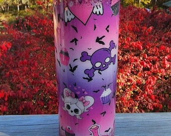 May include: A glittery, ombre-style tumbler in shades of pink, purple, and white, decorated with cartoon skulls, bats, and cupcakes. The design features a gradient effect, transitioning from pink to purple. The tumbler is cylindrical and appears to be made of stainless steel.