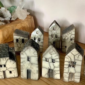 Könnte beinhalten: Eine Sammlung kleiner Keramikhäuser mit cremefarbenem Sockel und dunkelgrauen Dächern. Die Häuser haben ein strukturiertes, rissiges Aussehen und verfügen über kleine Fenster und Türen. Sie sind auf einer Holzoberfläche angeordnet.