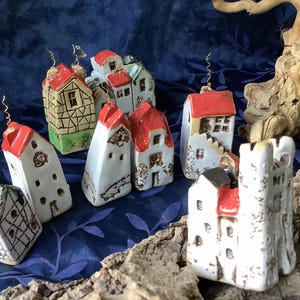 Könnte beinhalten: Eine Sammlung von Miniatur-Keramikhäusern mit roten, weißen und grünen Akzenten. Die Häuser haben unterschiedliche Designs, darunter Satteldächer und dekorative Details. Einige haben goldfarbene Drahtakzente.