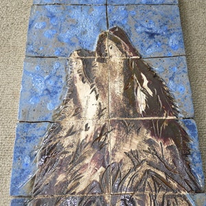 Pode incluir: Um mosaico de azulejos de cerâmica retratando um lobo uivando contra um céu azul. O lobo é renderizado em tons de marrom e branco, com textura detalhada. Os azulejos são dispostos para formar a imagem.