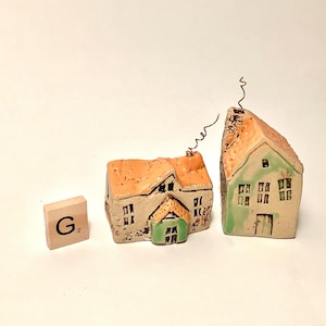 Puede incluir: Dos pequeñas casas de cerámica hechas a mano con detalles verdes y naranjas. Las casas están sobre una superficie blanca con una baldosa de madera que dice "G2" a su lado.