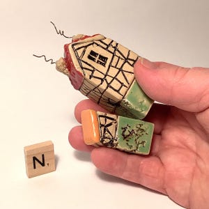 Puede incluir: Una pequeña casa de cerámica con un techo rojo, acentos verdes y naranjas y una ventana. La casa está rota en tres piezas y se sostiene en una mano. La letra "N" es visible en una baldosa de madera.