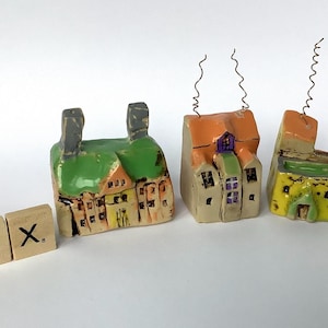 Op de afbeelding: Drie kleine keramische huisjes met groene daken en kleurrijke details. De huisjes zijn in een rij op een wit oppervlak gerangschikt. Het huisje links heeft twee schoorstenen, het middelste huisje heeft een schoorsteen met een draadspiraal, en het huisje rechts heeft een schoorsteen met een draadspiraal en een gele gevel.