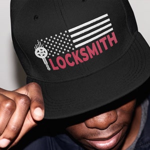 Op de afbeelding: Zwarte baseballpet met een rood-wit Amerikaans vlagontwerp en het woord "LOCKSMITH" in rode letters.