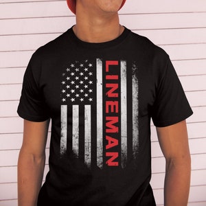 Puede incluir: Una camiseta negra con un diseño de bandera estadounidense desgastado. La palabra "LINEMAN" está impresa verticalmente en rojo en el lado derecho de la bandera.