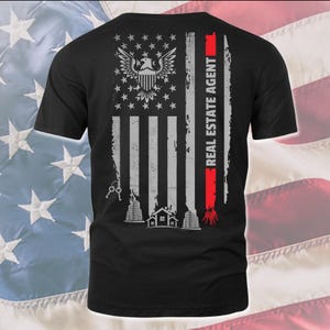 May include: Black t-shirt with a distressed American flag design. The flag is gray and white with a red stripe on the right side. The text "REAL ESTATE AGENT" is printed vertically on the red stripe. A gray eagle with outstretched wings is centered on the flag. The eagle is holding a key in its beak. There are gray buildings and a house at the bottom of the flag.