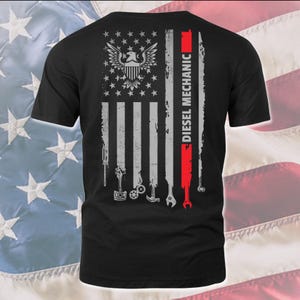 May include: Black t-shirt with a grey and white American flag design. The flag has tools and the text "Diesel Mechanic" on the red stripe.