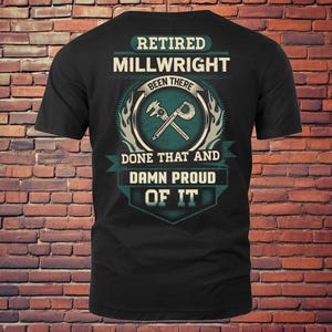 Op de afbeelding: Zwart t-shirt met de tekst "RETIRED MILLWRIGHT BEEN THERE DONE THAT AND DAMN PROUD OF IT". Het ontwerp bevat een cirkelvormig embleem met gereedschap en vlammen.