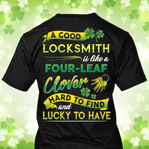 Puede incluir: Camiseta negra con un diseño gráfico verde y amarillo. El texto dice "A GOOD LOCKSMITH is like a FOUR-LEAF Clover HARD TO FIND and LUCKY TO HAVE." El diseño incluye tréboles y un juego de llaves.