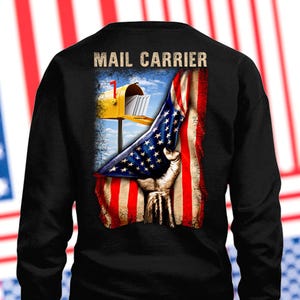 Könnte beinhalten: Schwarzes Sweatshirt mit einer Grafik einer gelben Postbox, einer amerikanischen Flagge und einer Hand, die die Flagge hält. Die Worte "MAIL CARRIER" sind oben aufgedruckt. Das Design hat eine verwitterte, patriotische Ästhetik.
