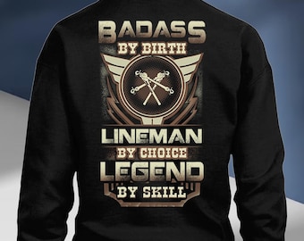 Op de afbeelding: Zwart sweatshirt met een grafisch ontwerp. Het ontwerp bevat de woorden "BADASS BY BIRTH", "LINEMAN BY CHOICE" en "LEGEND BY SKILL". De afbeelding bevat ook een cirkelvormig embleem met gekruiste gereedschappen en vleugels.