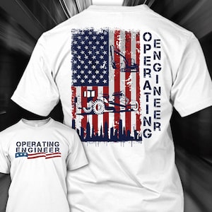 Puede incluir: Camiseta blanca con un diseño desgastado de la bandera estadounidense, vehículos de construcción y el texto "OPERATING ENGINEER". Una camiseta más pequeña con el mismo diseño es visible en primer plano.
